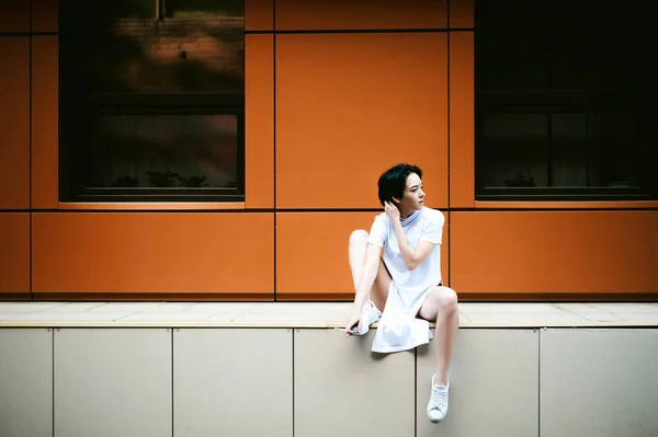 Jonge vrouw met de blote voeten gekleed in witte sneakers en een lang T-shirt, rookt een sigaret tegen de achtergrond van stadsgezichten. Straat mode — Stockfoto