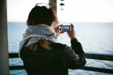 soğuk bir gün sahilde, bir atkı sarılı kadın telefonda bir fotoğraf alır