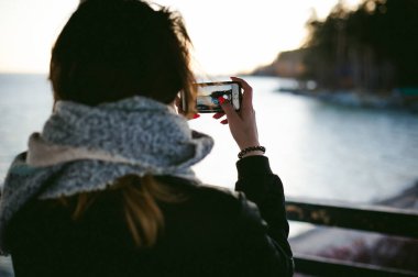 soğuk bir gün sahilde, bir atkı sarılı kadın telefonda bir fotoğraf alır