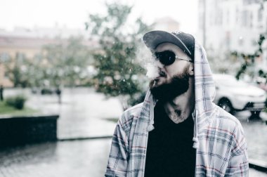 Kapüşonlu, kapüşonlu ve güneş gözlüklü sakallı genç adam şehirde serbestçe dolaşıyor ve bir e-sigara içiyor.