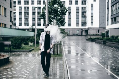 Kapüşonlu, kapüşonlu ve güneş gözlüklü sakallı genç adam şehirde serbestçe dolaşıyor ve bir e-sigara içiyor.