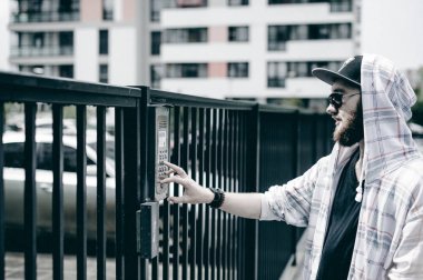 interkom aramak için çift sayı tuş takımındaki hood bir şapka ve güneş gözlüğü ile hafif kareli ceket içinde sakallı genç adam alır