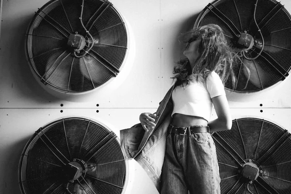 street portrait of a young attractive emotional girl with curly hair dressed in a trendy denim suit on the style. against the background of air conditioner ventilator systems. the wind in her hair