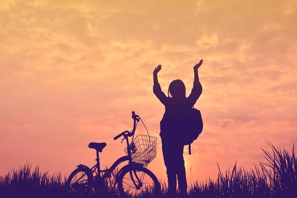 Silhueta de menina com bicicleta no campo de grama no pôr do sol céu — Fotografia de Stock