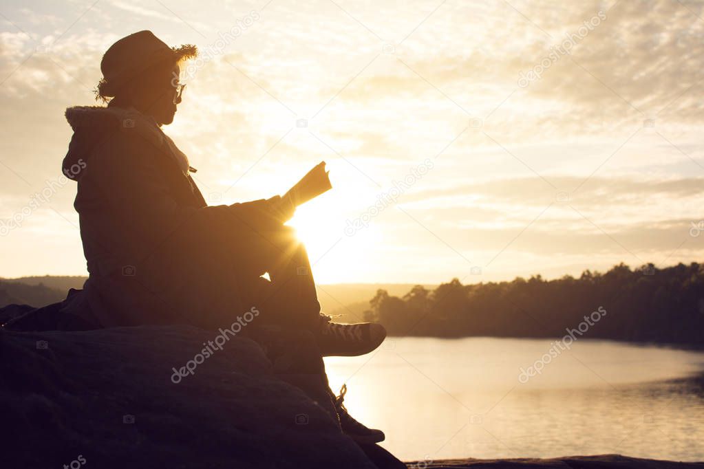 Silhouette of happy old women reading a book sitting on the rock near lake waiting for sunset relax time and holiday, color vintage style selective and soft focus