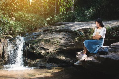 Asyalı kadınlar şelale orman arka plan, Relax zaman ve hobi yakınındaki kayaya oturan tatil, yumuşak odak, hippi tarzı tonunu bir kitap okuma