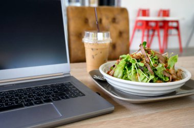 Baharatlı domuz salatası ve ahşap masada buzlu kahve ile modern laptop.