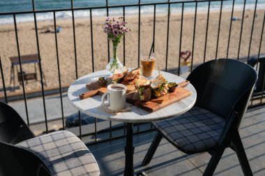 Sabahleyin kumsalın yanındaki terasta buzlu kahve ve çiçek süslemeli tavuklu sandviç.