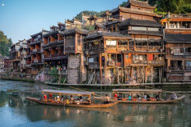 Feng Huang, Çin - Ağustos 2019: Fenghuang Eski Kasabası 'nın merkezinden akan Tuo nehrinin kıyısında inşa edilmiş eski tahta evlerin arasında seyir halindeki eski ahşap turist tekneleri