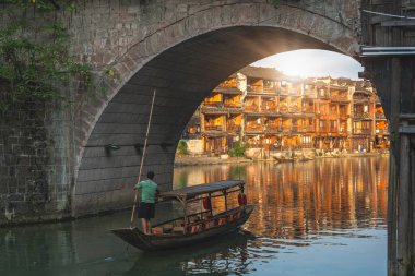 Feng Huang, China -  August 2019 :  Feng Huang, China - August 2019 : Old historic wooden tourist boat sailing under the iconic landmark arched bridge on the Tuo river, flowing through the centre of Fenghuang Old Town clipart