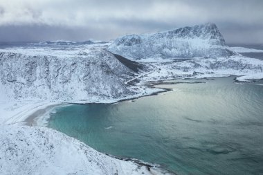 Kış mevsiminde Haukland Sahili 'ndeki Mannen Dağı' ndan Lofoten manzarası. Karların üzerindeki fırtınalı bulutlar dağları kapladı. Sakin plaj ve turkuaz deniz manzarası zirveden