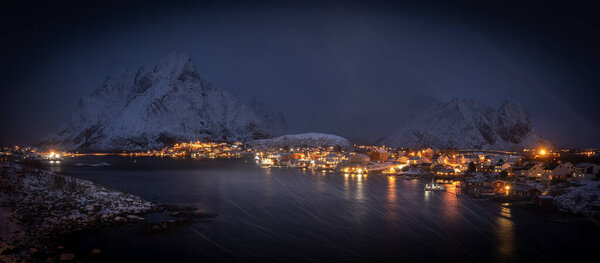 Night panorama of famous fishing village Reine, lofoten in heavy snowstorm. Blizzard and strong wind at harbor - in backgroud dark mountains covered with snow