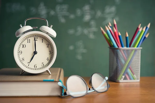 Συναγερμός ρολόι λευκό και πολύχρωμο μολύβι, Βιβλίο, Γυαλιά σε ξύλινο τραπέζι σε μαυροπίνακα φόντο στην τάξη — Φωτογραφία Αρχείου