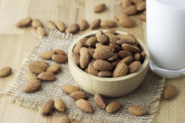 Çuval bezinde bir kase tahtanın içindeki badem tohumları ve masa tahtasında bir bardak süt.