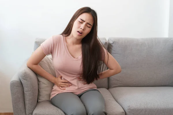 Femme asiatique avec menstruations et douleurs crampes menstruelles. jeunes femmes ayant douloureux assis sur le canapé à sa maison — Photo