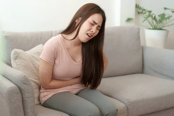 Mujer asiática con menstruación y calambres del período de dolor. mujeres jóvenes teniendo doloroso sentado en el sofá en su casa — Foto de Stock