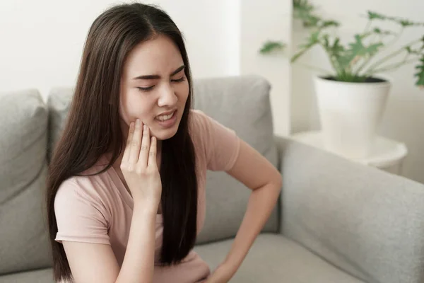 亚洲女人在家里坐在沙发上的时候牙疼 亚洲姑娘脸上的表情 概念牙痛及牙科药物 — 图库照片