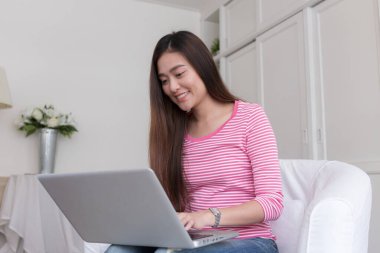 Asya 'lı genç ve güzel bir kız, oturma odasındaki kanepede otururken dizüstü bilgisayar kullanıyor, Asyalı kadınlar evde yazarken gülümsüyor.