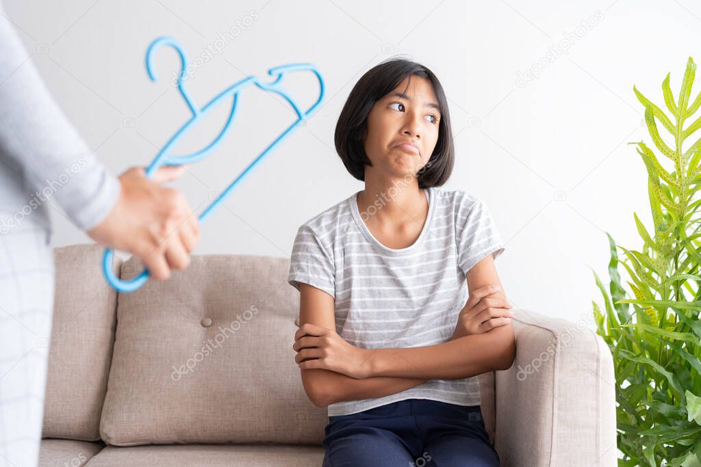 Mother is punishing her daughter by hitting with a hanger while sitting on the sofa because of bad behavior. The concept of family violence.