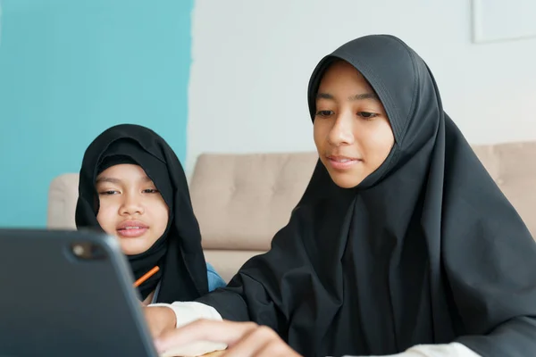 Two Muslim girl is studying online via the internet on tablet in living room at home, Asian elementary school children watching computer tablet. Concept of education at home