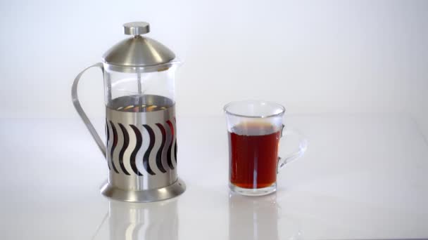 Tetera transparente con té y una taza con té caliente sobre fondo blanco — Vídeos de Stock