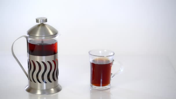 Tetera transparente con té y una taza con té caliente sobre fondo blanco — Vídeo de stock