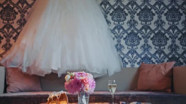 Vestido de novia colgando en la pared, ramo y zapatos están sobre la mesa — Vídeo de stock