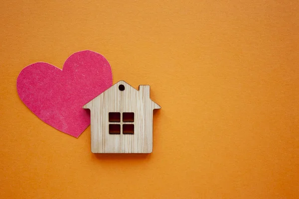 Love home, insurance and mortgage, marriage and valentine concept. Small wooden house toy and paper heart shape on orange background top view copyspace