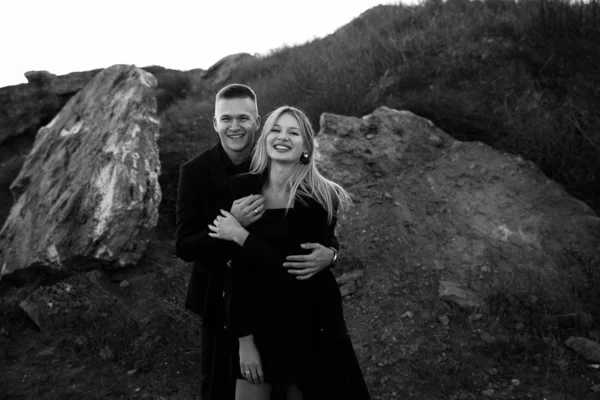 Retrato en blanco y negro de dos jóvenes amantes que se ríen al atardecer sobre fondo barranco. Pareja en ropa negra —  Fotos de Stock