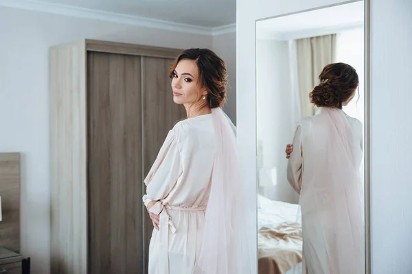 Beautiful bride is preparing for a meeting with her groom in the white room. Girl in wedding dress posing in a beige peignoir in front of the mirrow — Stock Photo, Image