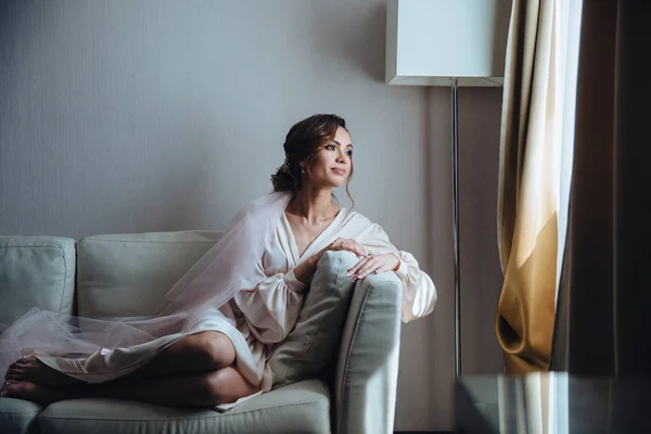 Mariée du matin, belle mariée attend le mariage assis sur le canapé dans la chambre blanche. Fille en robe de mariée devant la fenêtre attend le marié Images De Stock Libres De Droits