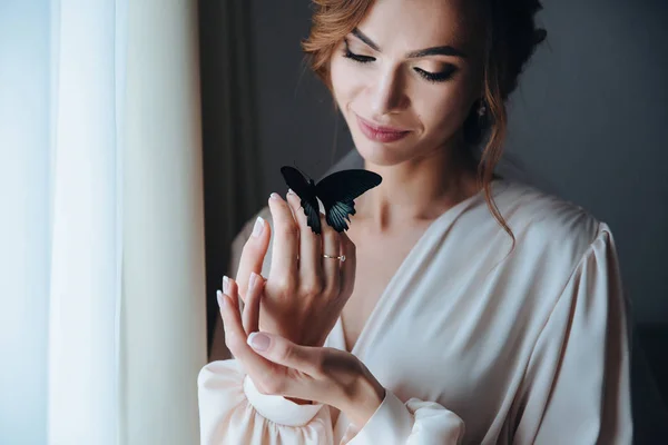 Primer plano retrato de una hermosa novia gentil en un peignoir beige con una mariposa con alas negras y azules se sienta en la mano de la novia —  Fotos de Stock