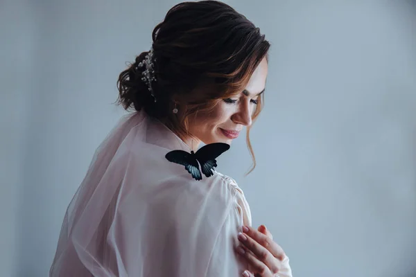 Een mooie zachte bruid in een beige peignoir met een vlinder met zwart-blauwe vleugels zit op de schouder van de bruid. Zijaanzicht — Stockfoto