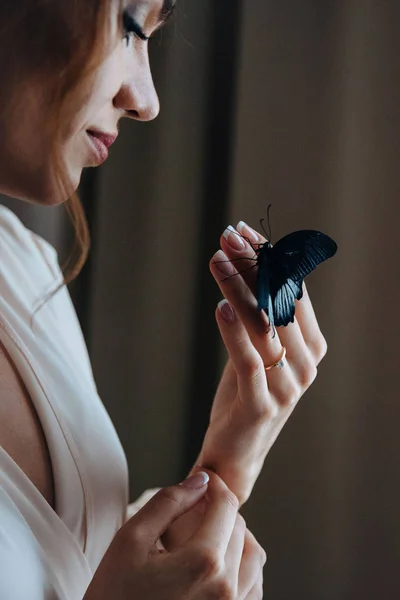 Close up portrait of A beautiful gentle bride in a beige peignoir with a butterfly with black and blue wings sits on the hand of the bride, she smiles. Side view — 스톡 사진
