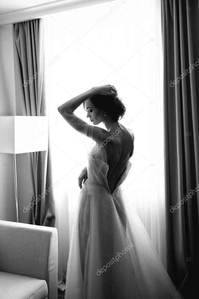 Black and white Portrait of the bride with open dress from the back on the window background. Beautiful neckline on the dress with sexy open back of the bride
