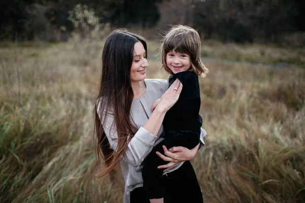Woman with son in her arms on dry grass field and forest background. Happy mom with long dark hair and her son — Stock Photo, Image