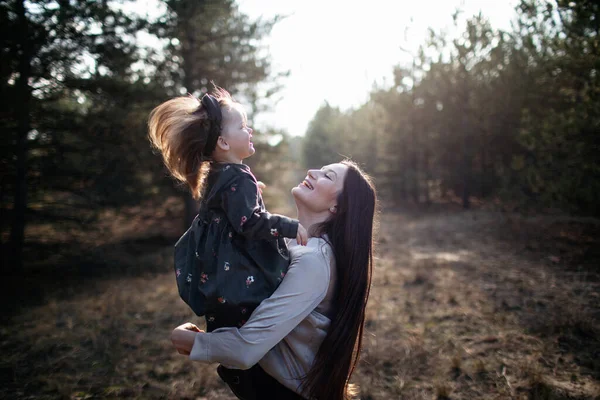 Joven hermosa mamá se da vuelta hija en sus brazos en el fondo del bosque en la puesta del sol. Riendo mamá y su hija. La paternidad. Mamá abraza hija —  Fotos de Stock