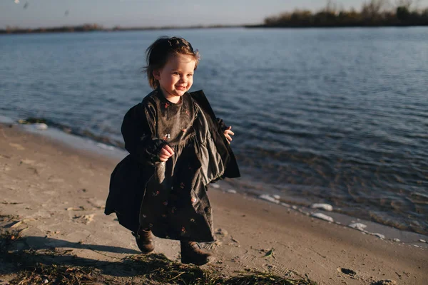 Niño caminando en la playa con ropa oscura. Cálido día de otoño. Una chica solitaria en la orilla del río al atardecer. Emociones de niños . —  Fotos de Stock