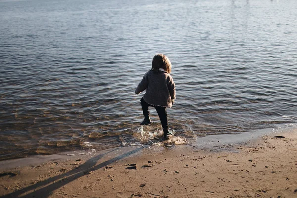 Kind läuft im Herbst im Wasser am Flussufer. Rückseite. warmer Herbsttag. Ungehorsam. ein einsames Kind am Ufer des Flusses bei Sonnenuntergang. Emotionen. — Stockfoto