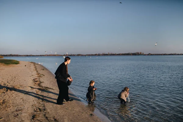 Enfants et maman courant dans l'eau sur la rive de la rivière en automne. Le garçon boit de l'eau de rivière. Désobéissance. Bonne famille au bord de la rivière Au coucher du soleil . Photo De Stock