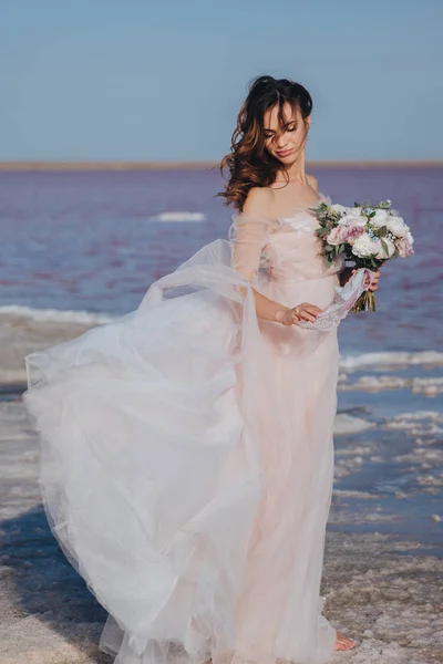 Sensuele bruid in een trouwjurk aan de kust met de wind in haar haar en vliegende jurk. Vooraanzicht. — Stockfoto