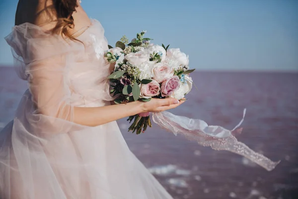 Gyengéd menyasszony elefántcsont menyasszonyi ruhában a tengerparton, a csokorral a kezében a tenger hátterében. Visszanézés. — Stock Fotó