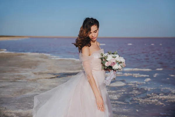 Sensuele bruid in een trouwjurk aan de kust met een boeket en de wind in haar haar. vooraanzicht. — Stockfoto