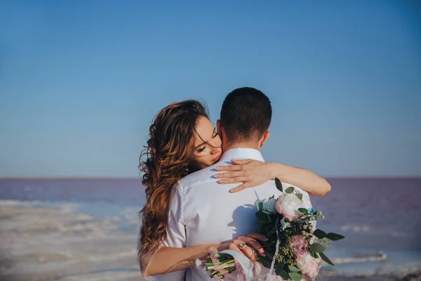 Belle mariée dans une robe de mariée sur le bord de la mer avec un bouquet serrant son marié. Vue arrière du marié — Photo