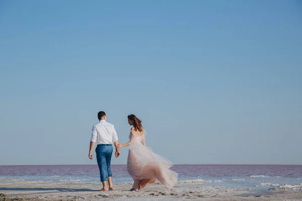 Pasgetrouwde echtparen die hun handen vasthouden en langs de kust lopen. Bruid in ivoor vliegende jurk. Achteraanzicht — Stockfoto