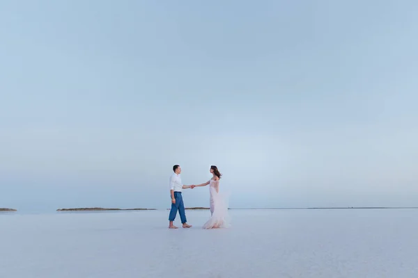 Mooie jonge pasgetrouwden aan de kust, de bruidegom hand in hand met de bruid op het witte zand. achteraanzicht. — Stockfoto