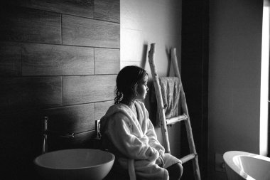 Pretty little girl in a white unicorn bathrobes sitting after having bath with wet hair and looking to the window