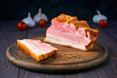 Tütsülenmiş domuz göbeği dilimlenmiş şerit ve tahtadan kesme tahtasının üzerinde tahıl ekmeği. Sebze ve baharatlar: domates, sarımsak ve kişniş. Kırsal biçim.