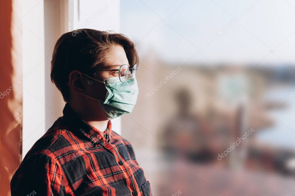 A girl in a protective mask from covid-19 stands at the window and looks at the nature. The concept of a coronavirus pandemic that has hit the whole world. stays at home. Copy space.