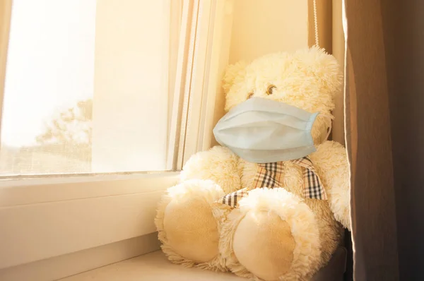 Epidemia koronawirusa, COVID-19.Smutny niedźwiedź siedzi w masce medycznej na parapecie i wygląda przez okno. Koncepcja światowej izolacji spowodowanej pandemią. Zostań w domu.. — Zdjęcie stockowe